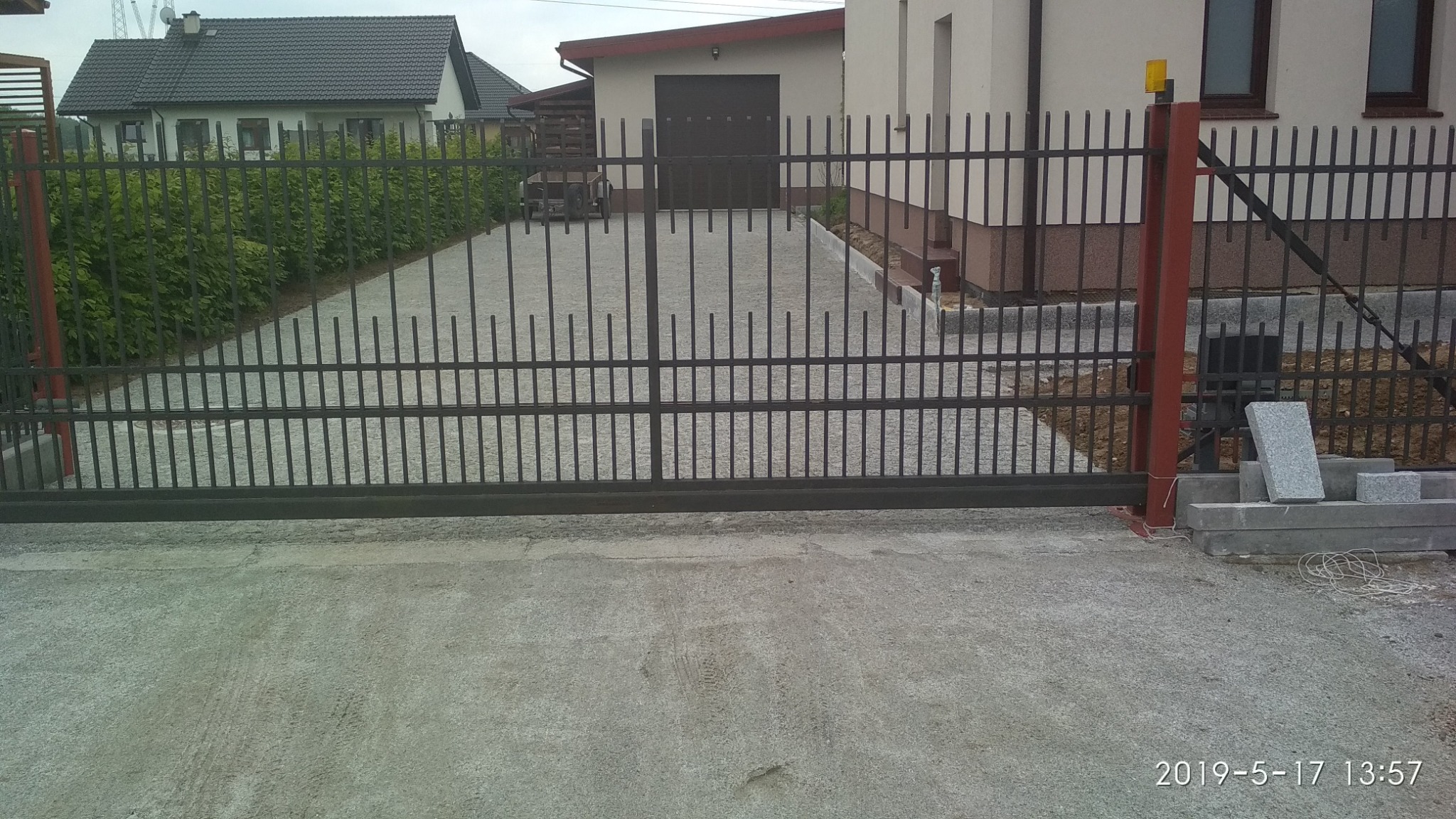 Nowoczesna, przesuwna brama wjazdowa z metalowymi prętami, widok na podjazd z betonową nawierzchnią oraz fragment domu w tle. Na pierwszym planie ułożone obrzeża z szarej kostki.