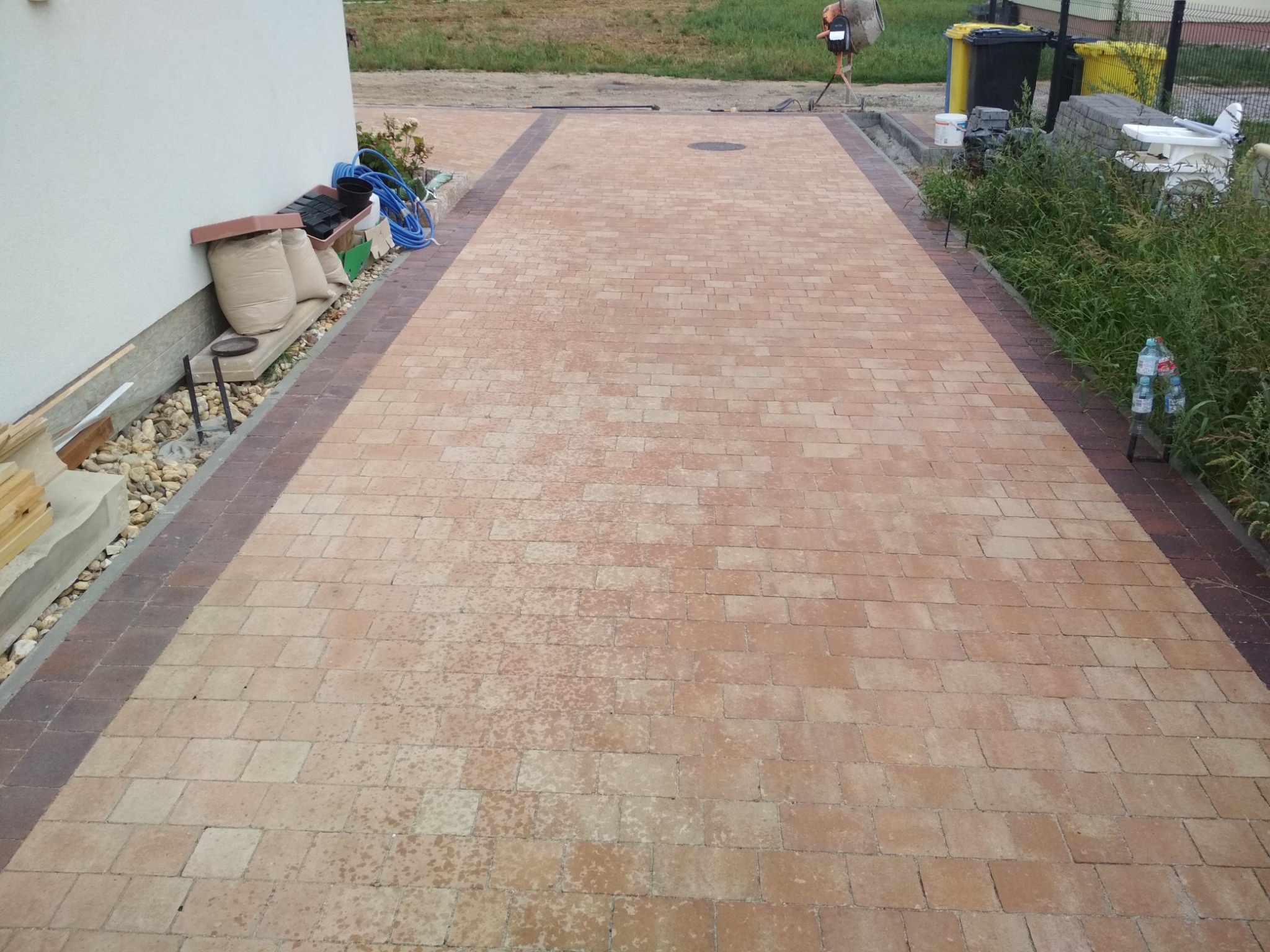 Nowo ułożona, beżowo-brązowa kostka brukowa na podjeździe, z widocznymi workami z materiałem budowlanym przy ścianie budynku i betoniarką w tle.