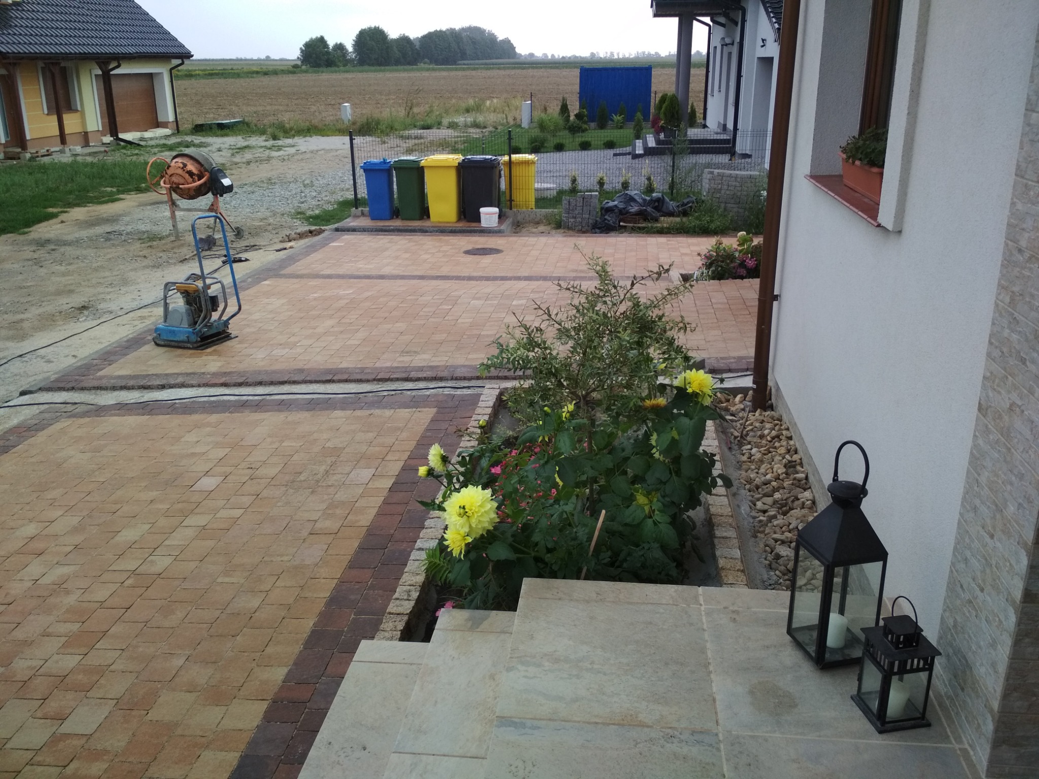 Ułożona kostka brukowa w kolorze beżu i brązu, obrzeże, widoczna zagęszczarka, rabata z kwiatami i schody wejściowe do domu z lampionami.