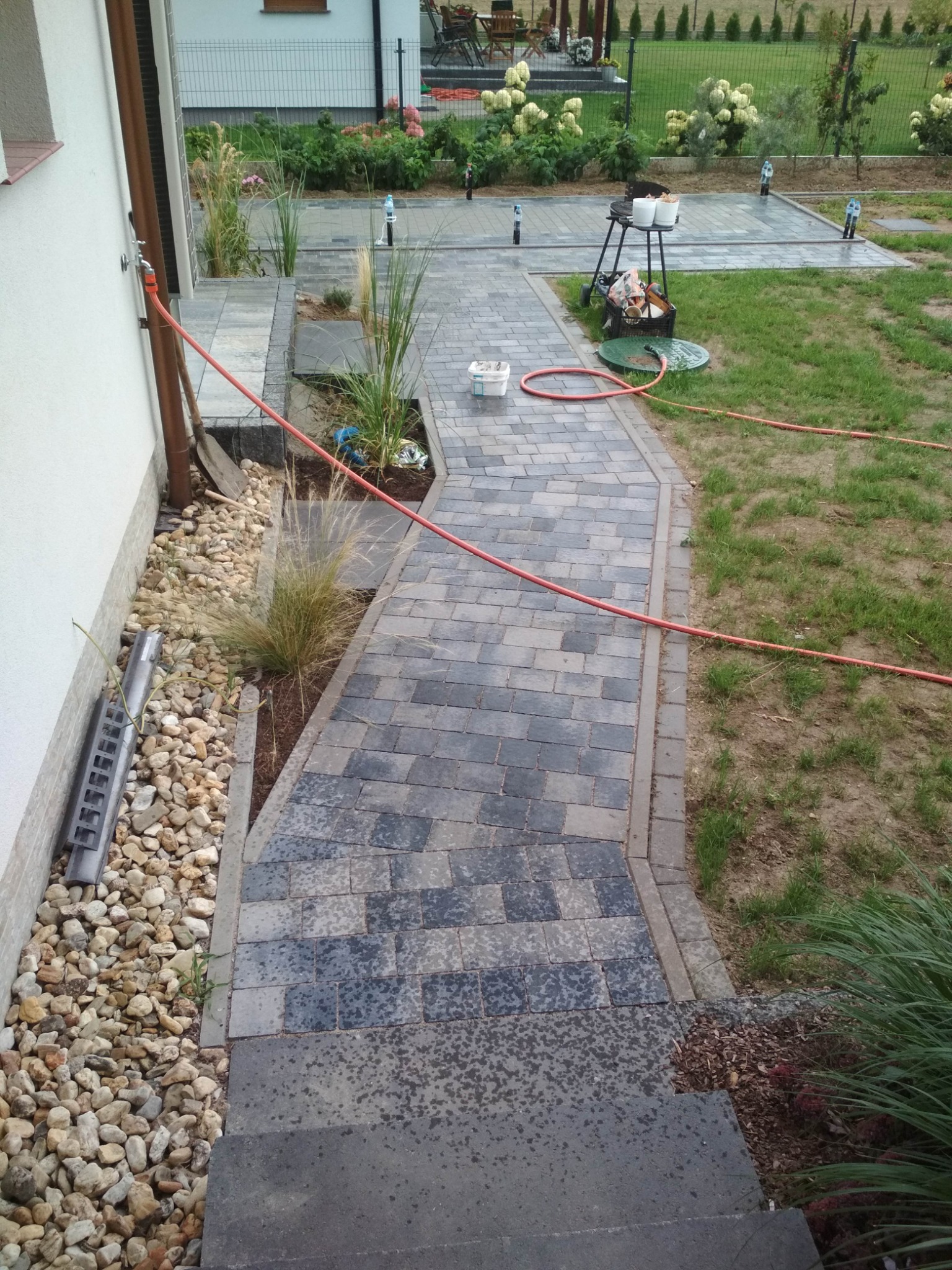 Ścieżka z kostki brukowej prowadząca od schodów wzdłuż domu w Męcince, z widoczną instalacją nawadniającą i elementami dekoracyjnymi z kamienia i roślin.