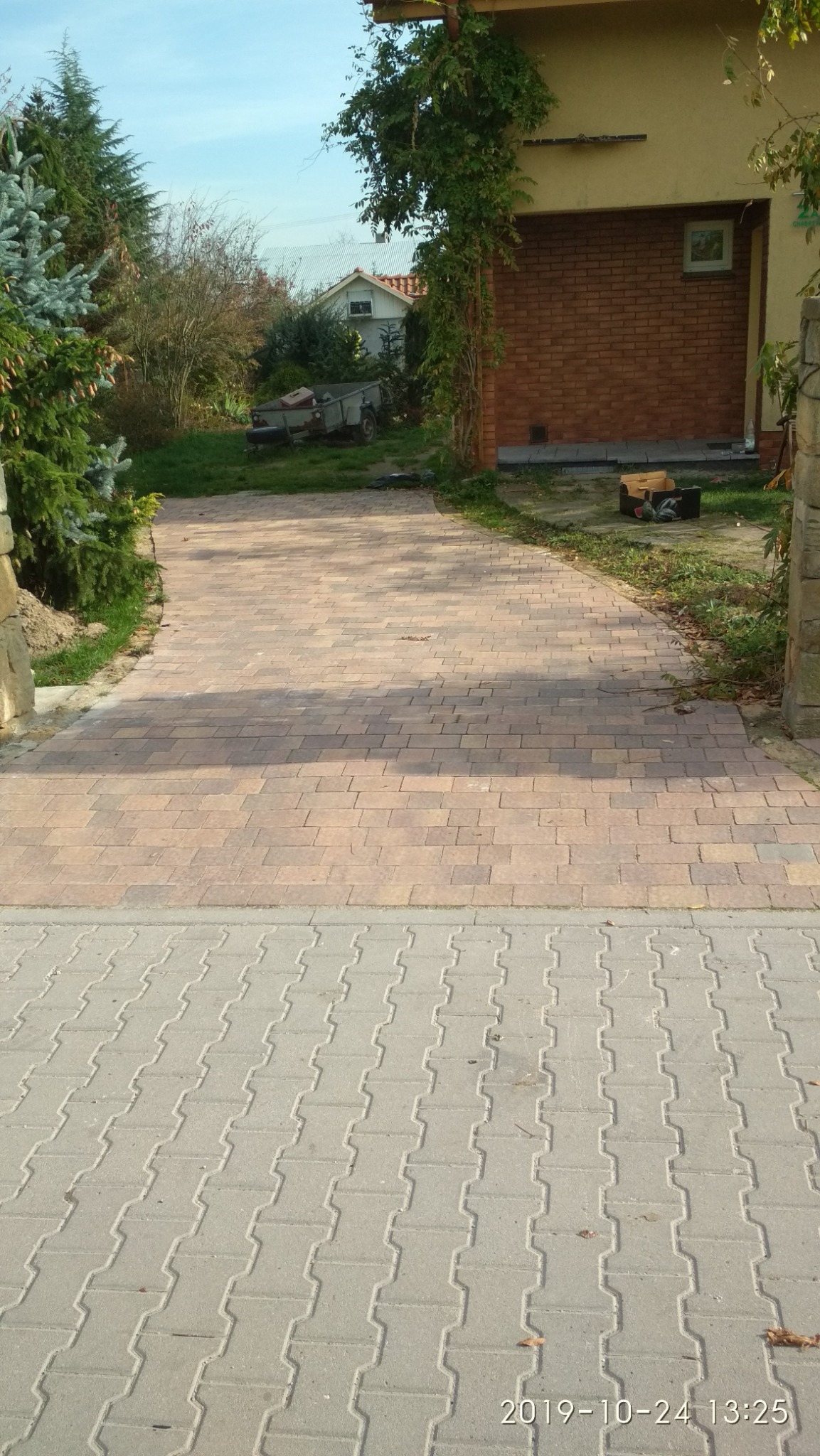 Szeroki podjazd wyłożony dwoma rodzajami kostki: szarą w kształcie litery H i czerwonobrązową prowadzący do budynku z cegły porośniętego bluszczem.