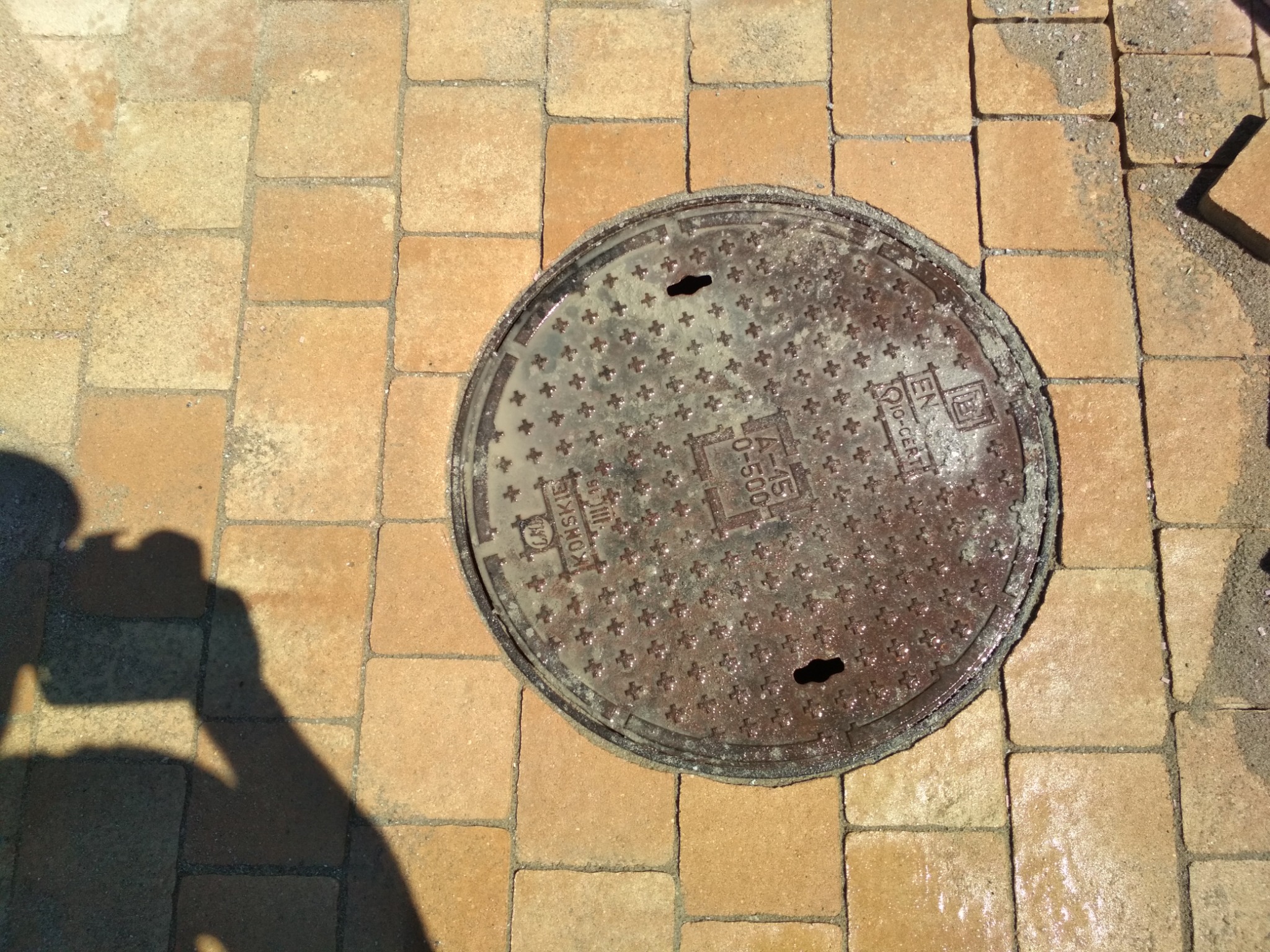 Pomarańczowa kostka brukowa ułożona wokół włazu kanalizacyjnego z cieniem osoby robiącej zdjęcie.