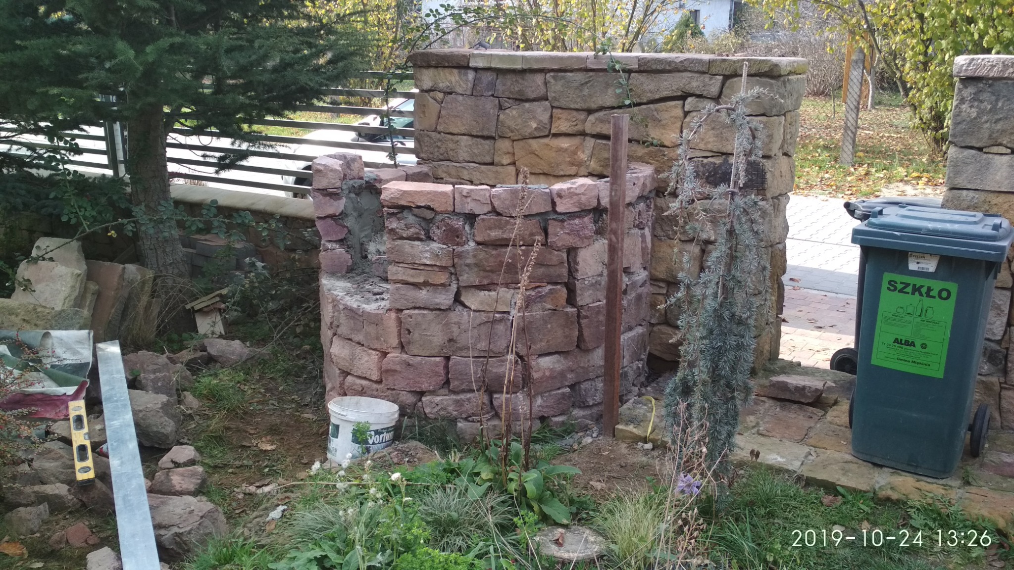 Konstrukcja z kamienia w trakcie budowy, widoczne elementy muru, poziomica, wiadro z zaprawą i pojemnik na szkło w ogrodzie.