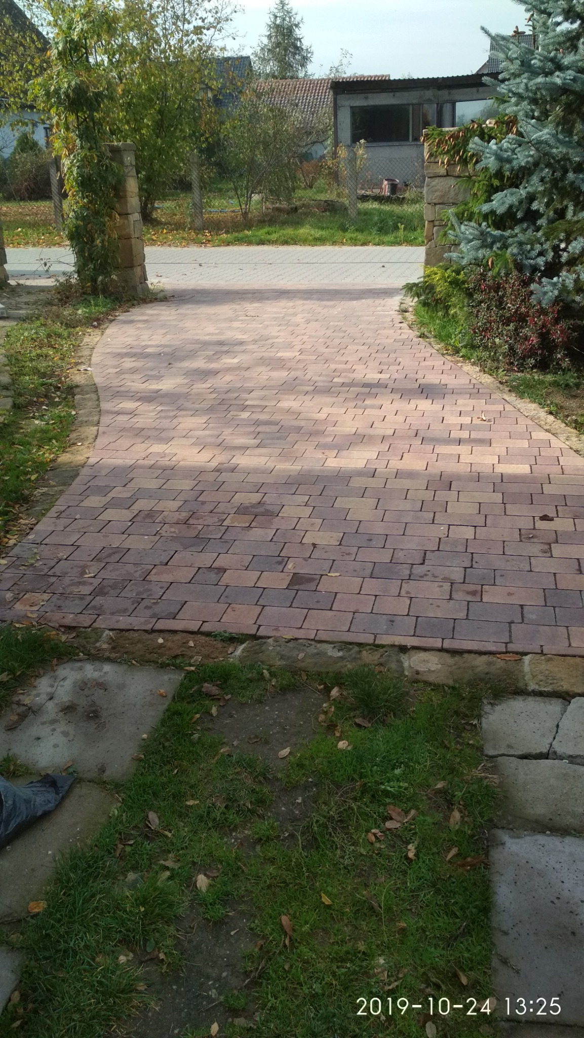 Ułożona ścieżka z kostki w odcieniach brązu i czerwieni prowadząca między kamiennymi słupkami porośniętymi zielenią, otoczona trawnikiem i drzewami.