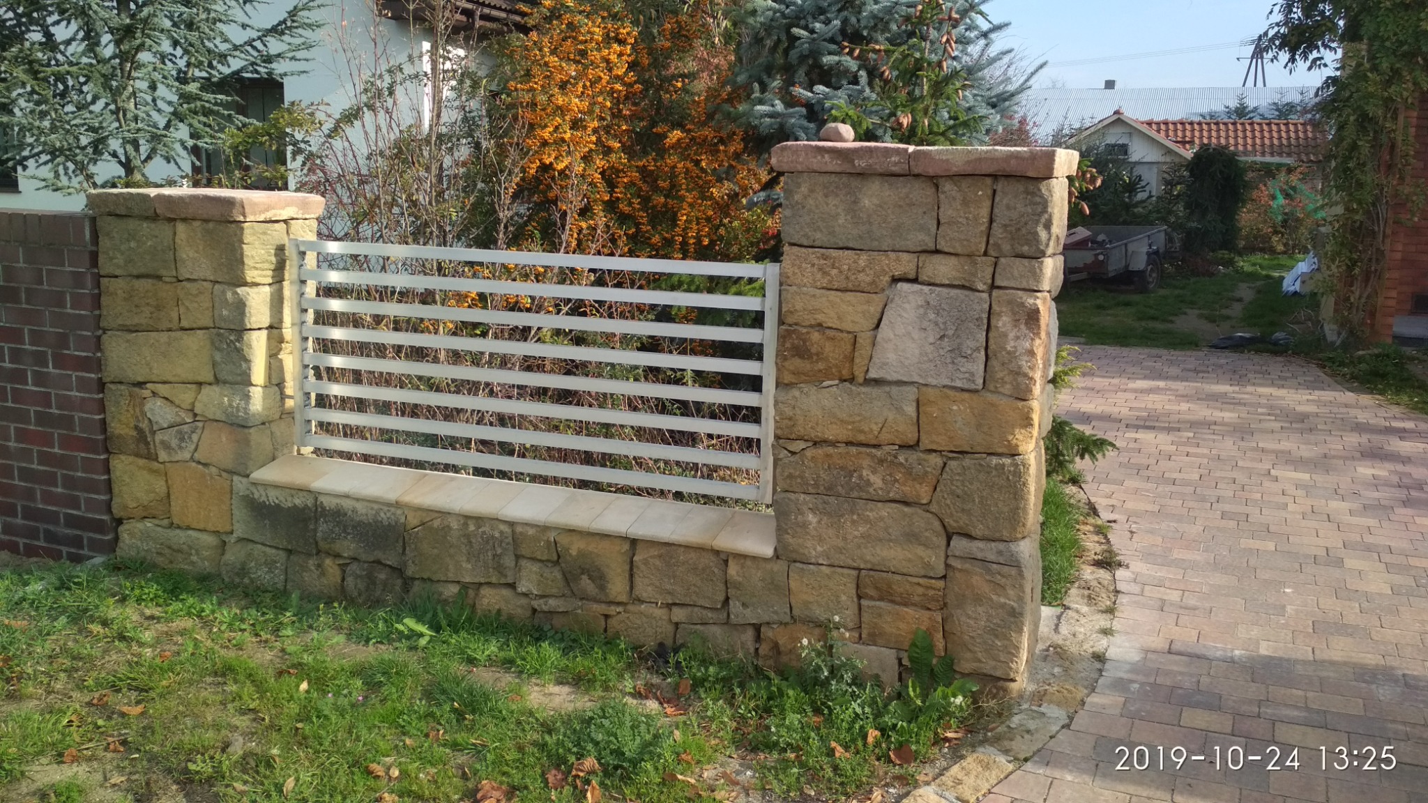 Murowane słupki z kamienia piaskowca połączone metalowym ogrodzeniem poziomym, otoczone zielenią i brukowanym podjazdem.