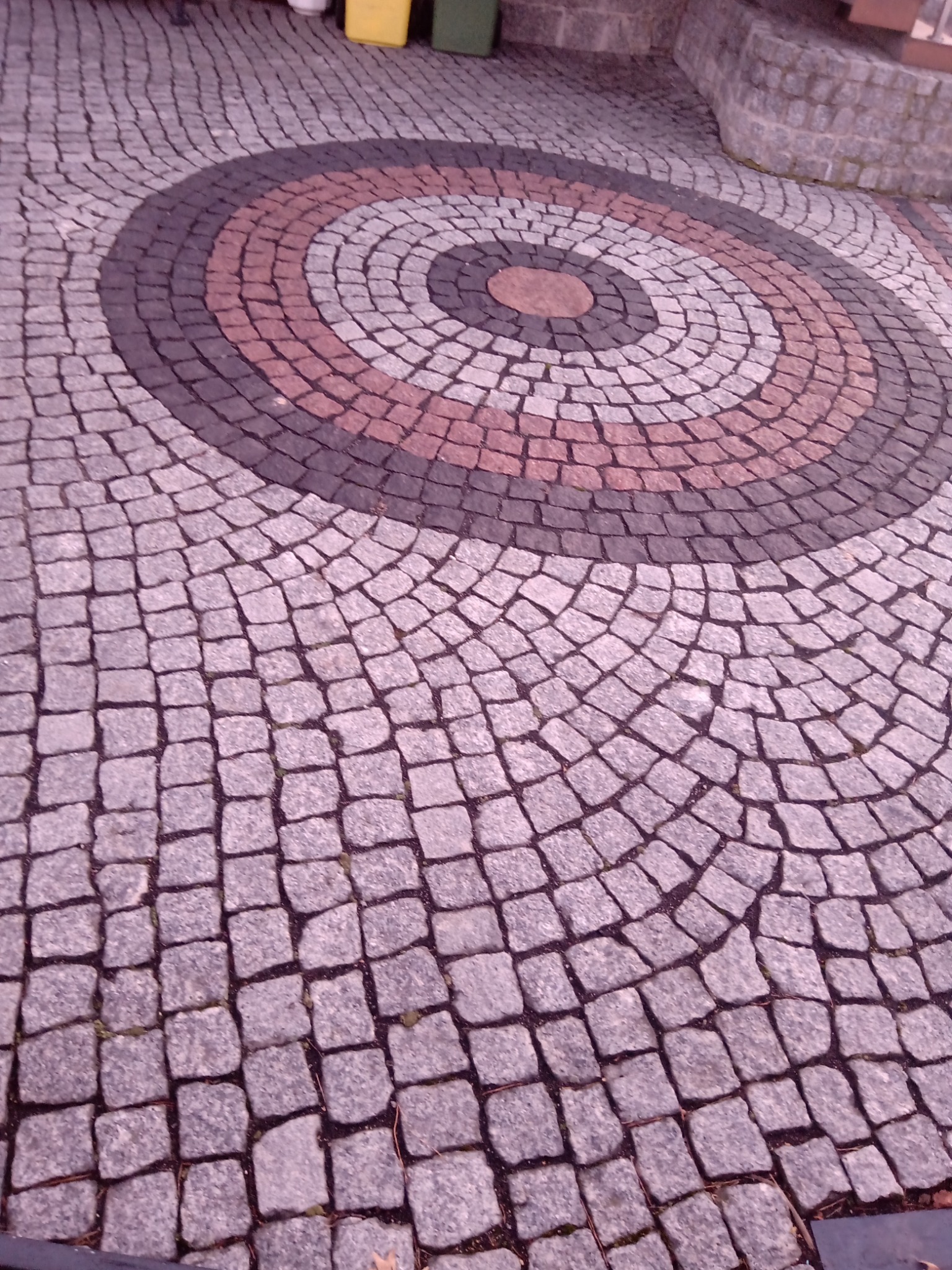 Wzór z kostki granitowej w kształcie okręgu, z użyciem szarej, czarnej i czerwonej kostki, tworzący dekoracyjny element na podwórku.