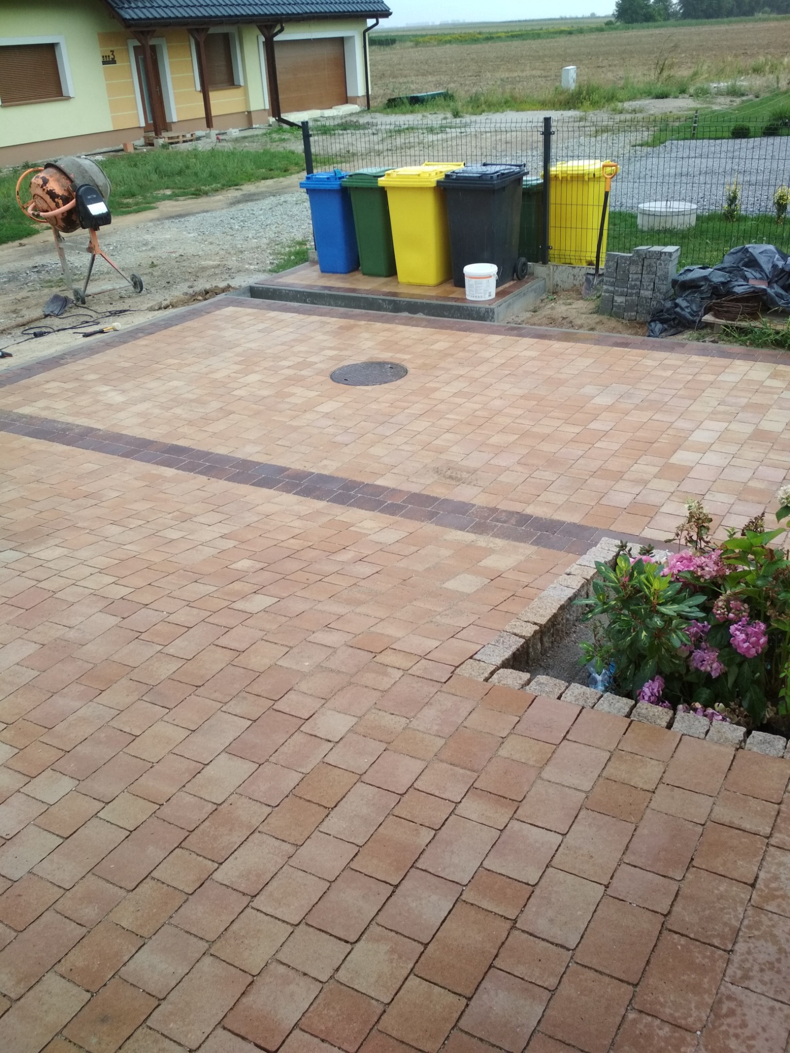 Nowo ułożona, beżowo-brązowa kostka brukowa na podwórku z ciemniejszym pasem kostki, w tle dom, pojemniki na śmieci i betoniarka.