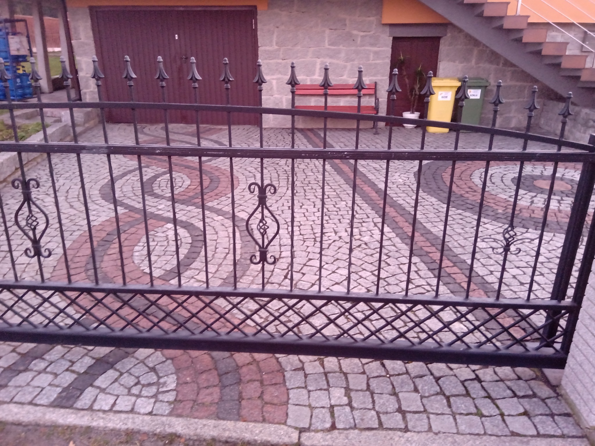 Wzór z kostki brukowej w odcieniach szarości i różu, widoczny przez czarne, metalowe ogrodzenie posesji z dekoracyjnymi elementami, w tle brama garażowa i ławka.
