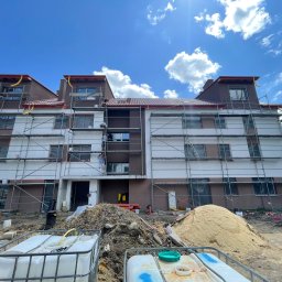 AGARPBUD Sp. z o.o. - Wielopiętrowy budynek w trakcie prac wykończeniowych elewacji, widoczne rusztowania, hałdy piasku i ziemi, oraz pojemniki z wodą na pierwszym planie.