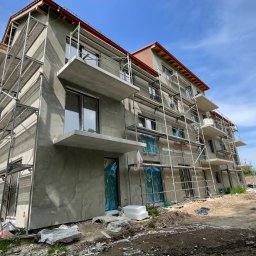 AGARPBUD Sp. z o.o. - Budynek wielorodzinny w trakcie prac wykończeniowych elewacji, widoczne rusztowania, surowa szara struktura tynku, okna zabezpieczone folią, balkony w stanie surowym.