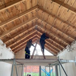 AGARPBUD Sp. z o.o. - Dwaj pracownicy na rusztowaniu i drabinie montują drewnianą konstrukcję dachu z widocznymi metalowymi łącznikami, w tle okno z niebieską taśmą zabezpieczającą.