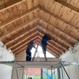 Dwaj pracownicy na rusztowaniu i drabinie montują drewnianą konstrukcję dachu z widocznymi metalowymi łącznikami, w tle okno z niebieską taśmą zabezpieczającą.