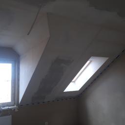 AGARPBUD Sp. z o.o. - Poddasze w trakcie prac wykończeniowych: ściany i skośny sufit pokryte świeżą warstwą gładzi, zabezpieczone taśmą malarską przy oknie dachowym i ścianie szczytowej z oknem, widoczny grzejnik...