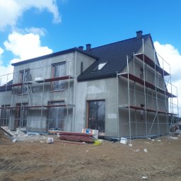 AGARPBUD Sp. z o.o. - Dwukondygnacyjny dom w trakcie ocieplania elewacji, z widocznymi rusztowaniami, oknami zabezpieczonymi folią i stertą materiałów budowlanych na ziemi.