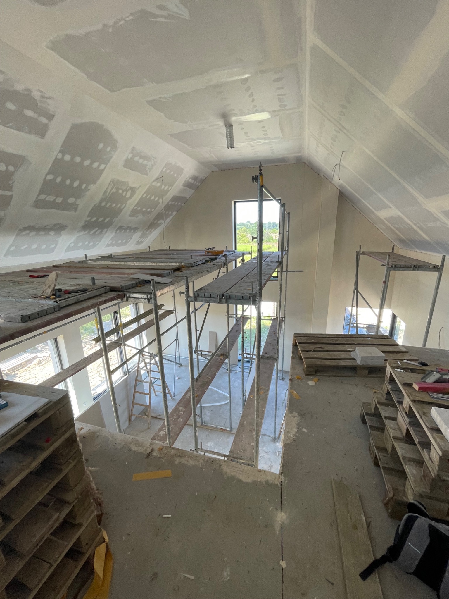 Poddasze w trakcie prac wykończeniowych: widoczne rusztowania, częściowo wykończone ściany i sufit z płyt kartonowo-gipsowych, szpachlowane łączenia, palety z materiałami budowlanymi.