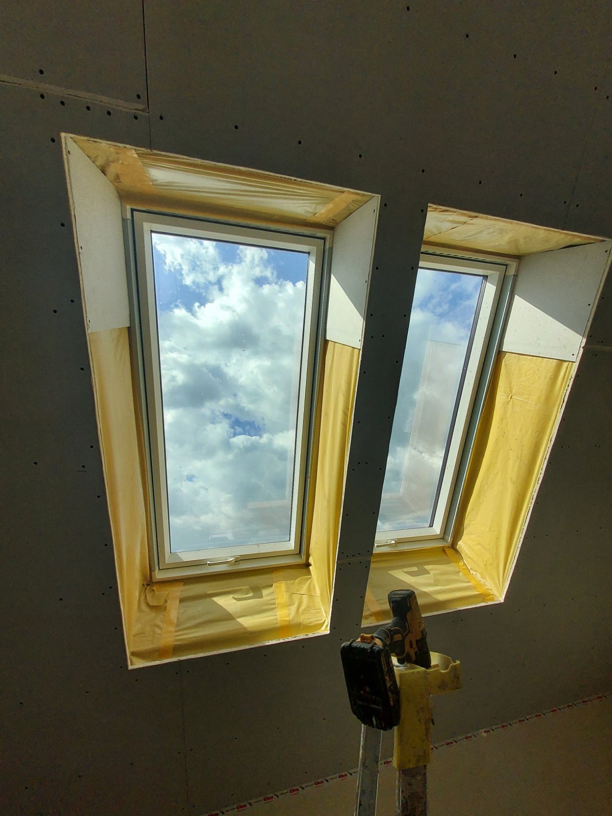 Dwa okna dachowe z widokiem na niebo z chmurami, zamontowane w skośnym suficie z płyt kartonowo-gipsowych, zabezpieczone żółtą folią ochronną, widoczny wiertarko-wkrętak na drabinie.