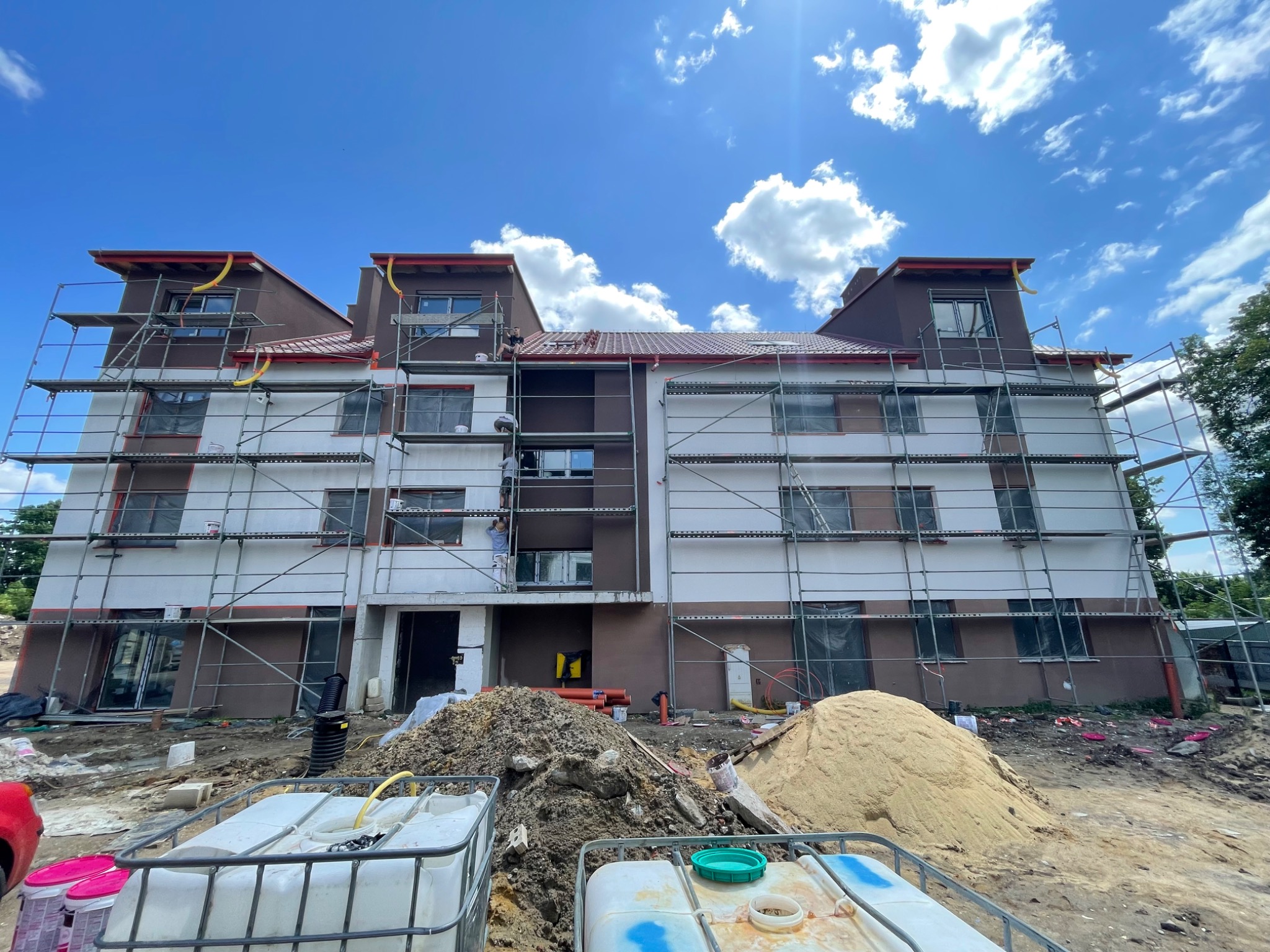 Wielopiętrowy budynek w trakcie prac wykończeniowych elewacji, widoczne rusztowania, hałdy piasku i ziemi, oraz pojemniki z wodą na pierwszym planie.