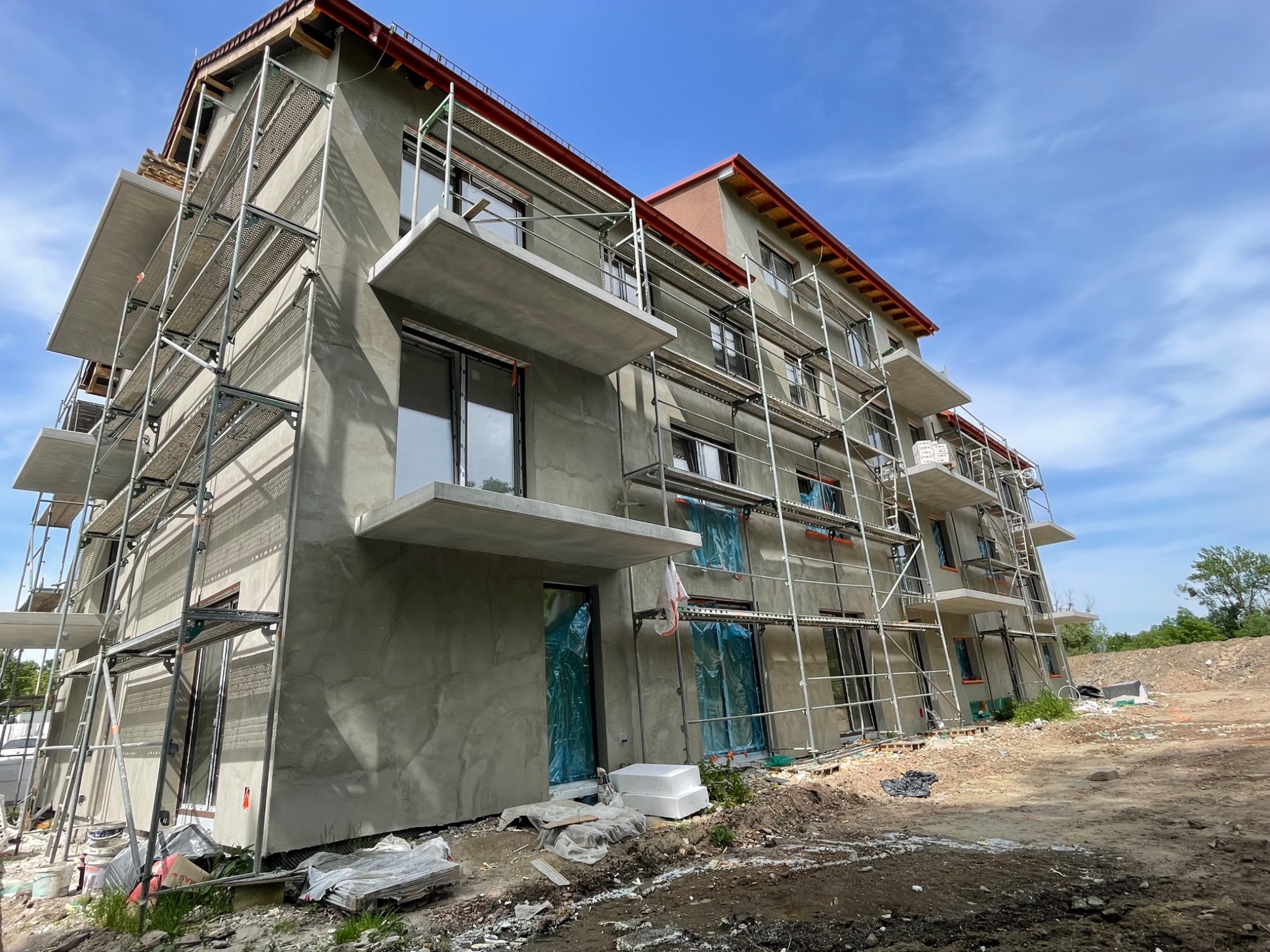 Budynek wielorodzinny w trakcie prac wykończeniowych elewacji, widoczne rusztowania, surowa szara struktura tynku, okna zabezpieczone folią, balkony w stanie surowym.