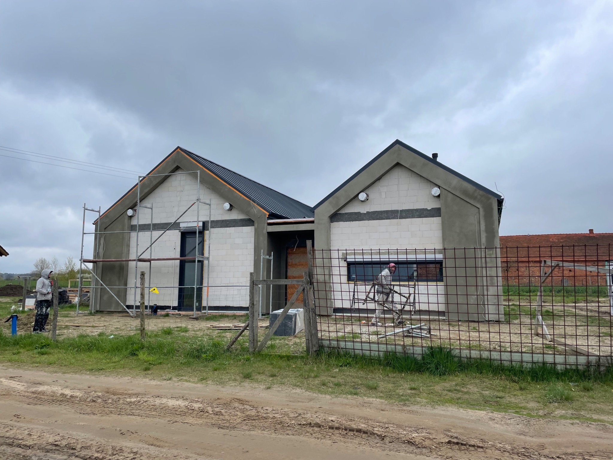 Surowy stan budynku jednorodzinnego w trakcie prac wykończeniowych elewacji, widoczne rusztowania, ocieplenie oraz pracownicy w kombinezonach ochronnych. Budynek z białego bloczka, z ciemnym dachem...