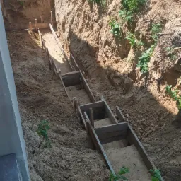 Wykopany rów z drewnianym szalunkiem przygotowany pod fundament, widoczne ślady ziemi na szalunku i roślinność na krawędzi wykopu.