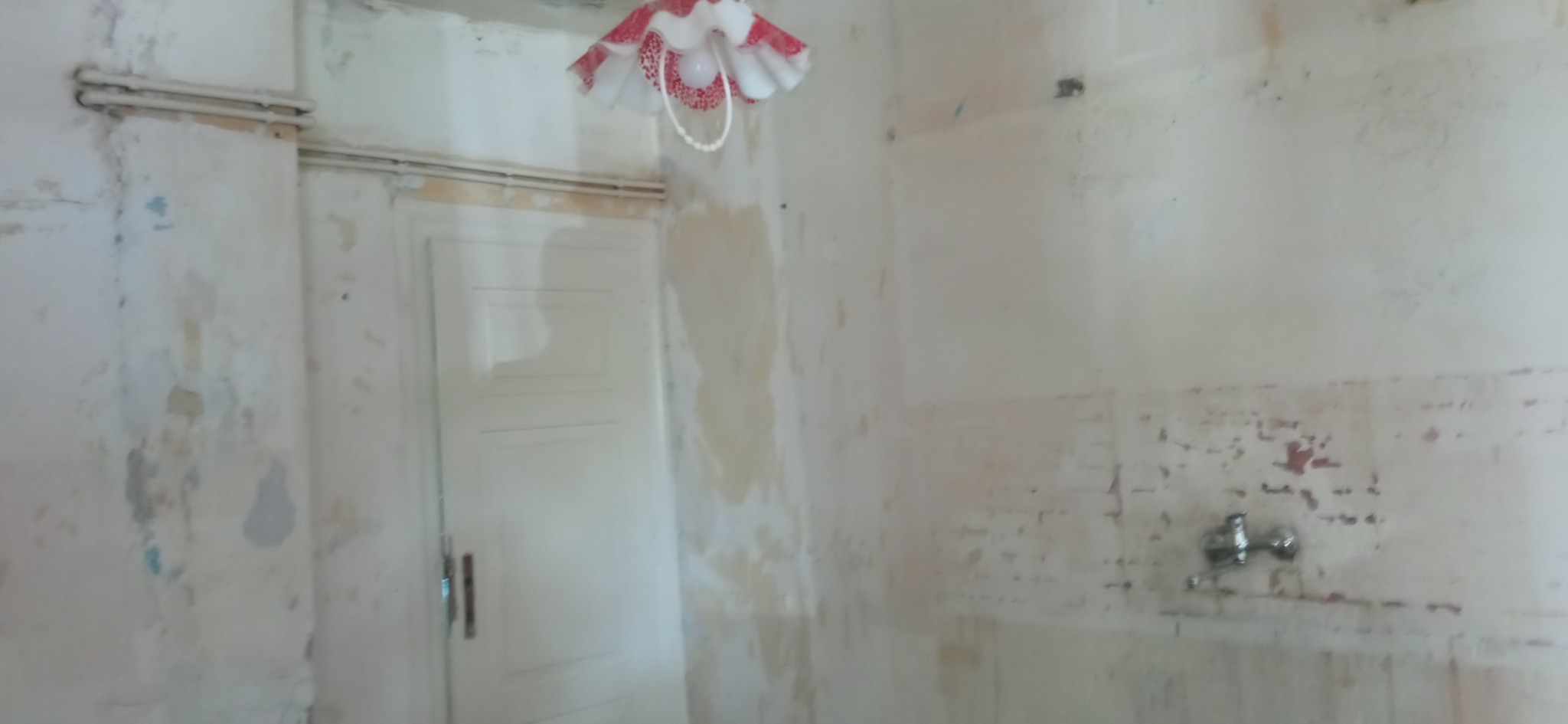 Zniszczone wnętrze łazienki z odpadającą farbą, widoczne ślady po remoncie, stara armatura i lampa z abażurem w kwiaty.