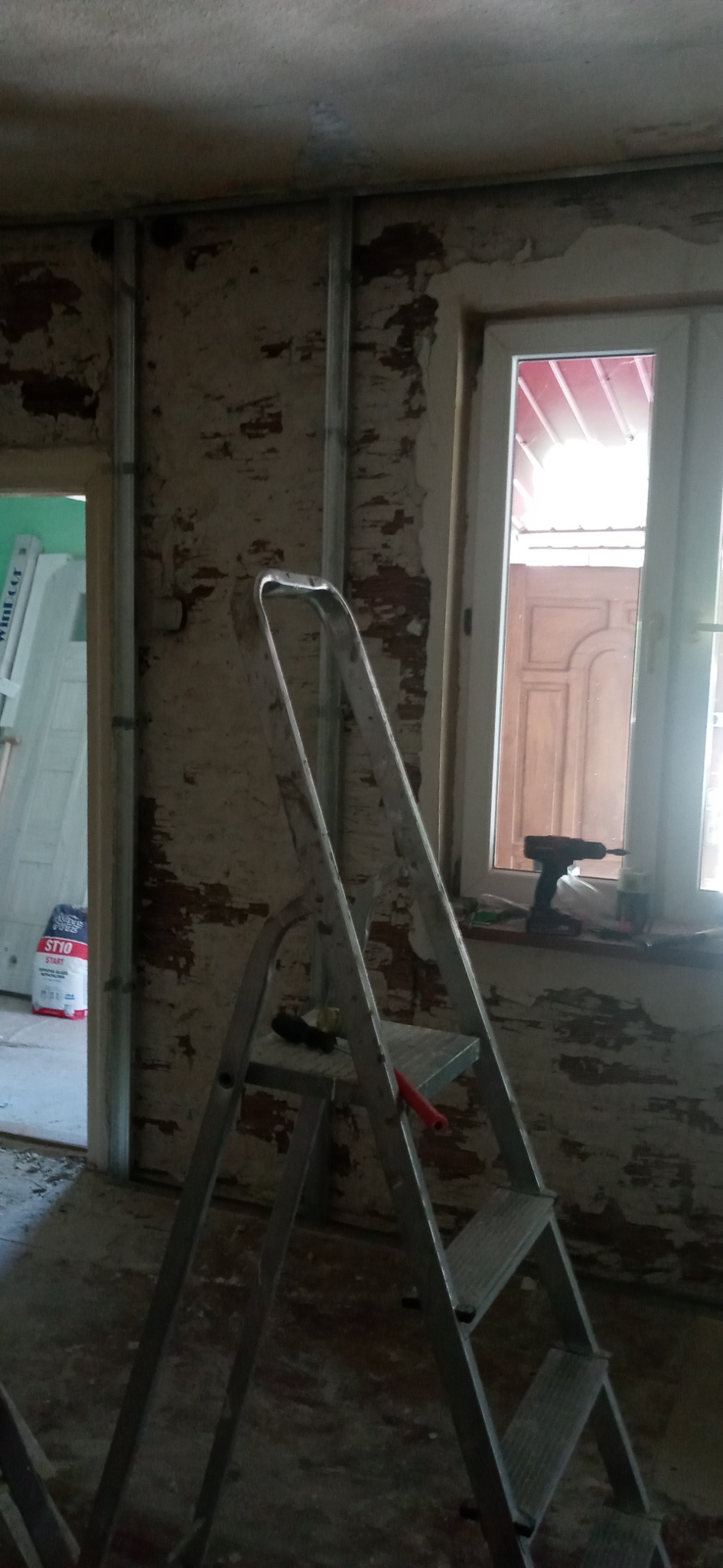 Ściana w trakcie remontu z widocznymi fragmentami starego tynku, metalowe profile pionowe, drabina aluminiowa, wiertarka akumulatorowa na parapecie, worek gładzi szpachlowej w tle.