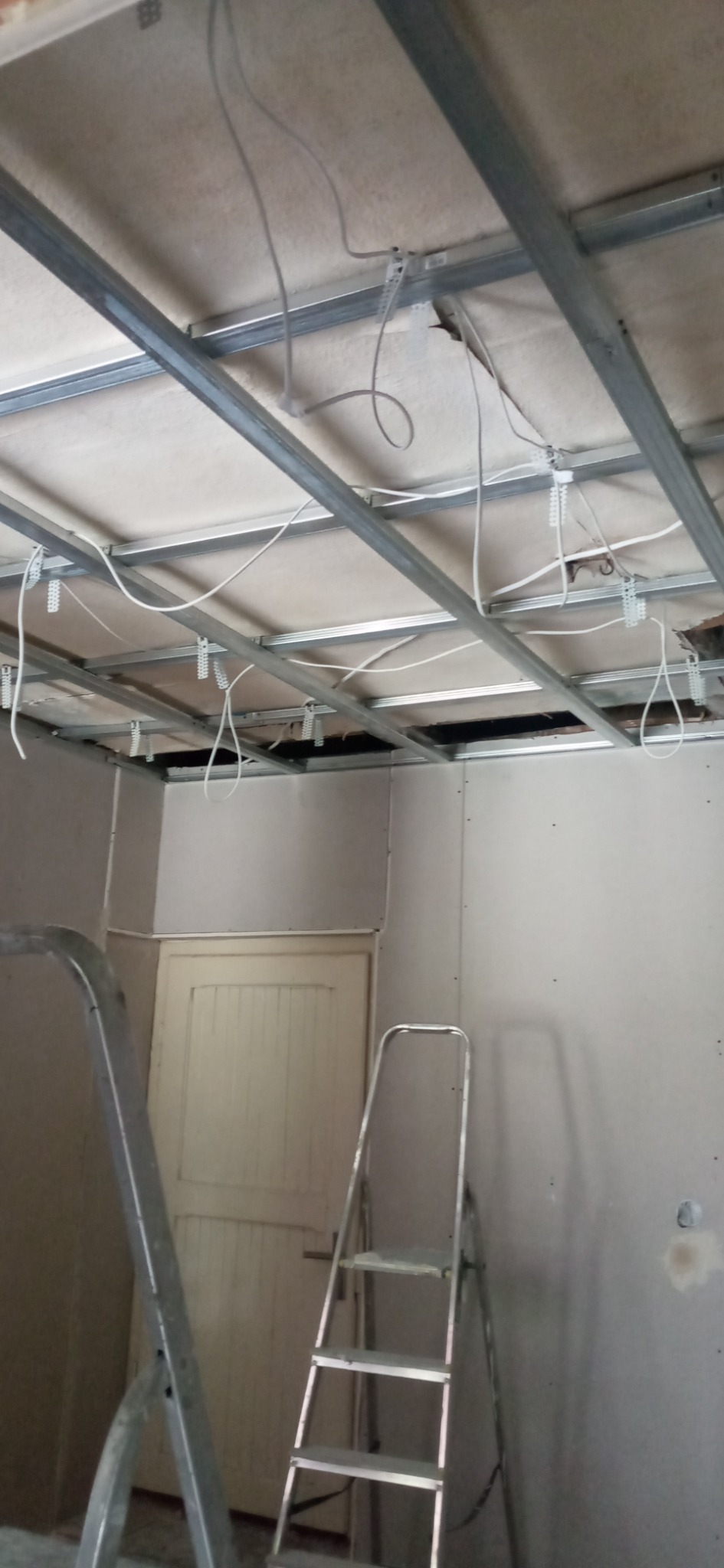 Montaż sufitu podwieszanego z płyt kartonowo-gipsowych, widoczne profile metalowe, instalacja elektryczna i drabiny w pomieszczeniu przed wykończeniem.