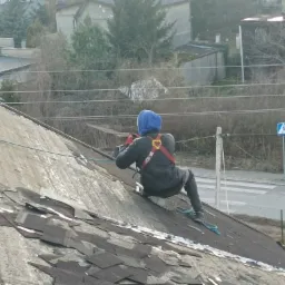 Pracownik w uprzęży zabezpieczającej na stromym dachu krytym łupkiem, w trakcie prac dekarskich, widok z góry na tle zabudowań i ulicy z przejściem dla pieszych.