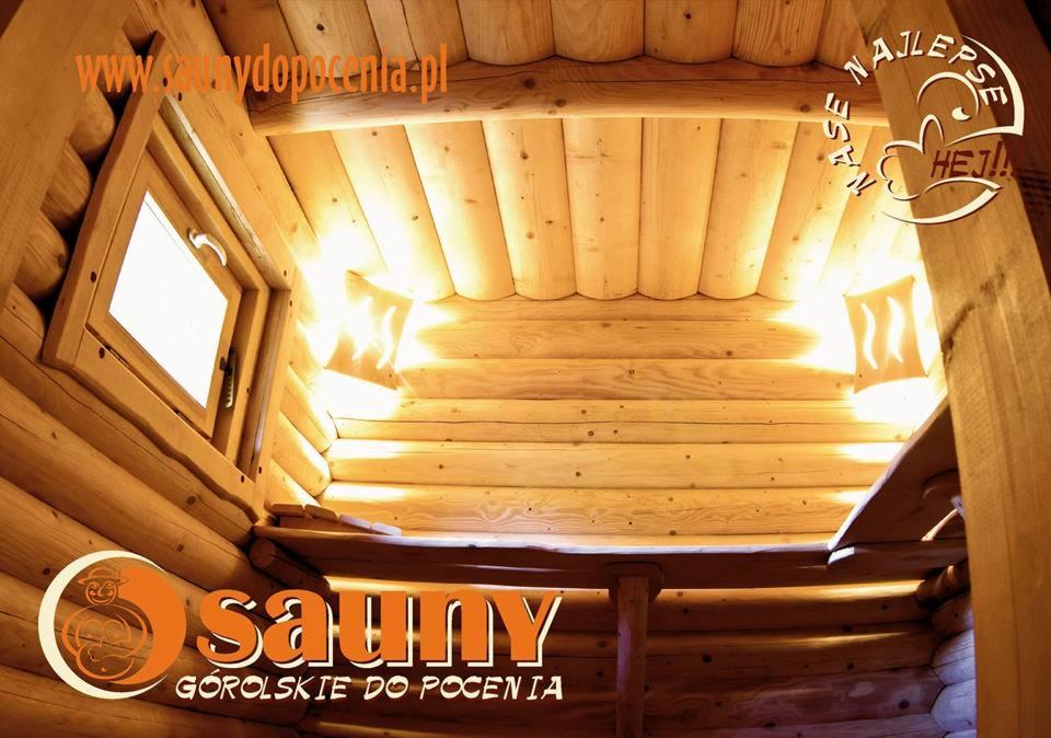 Wnętrze sauny góralskiej z bali, oświetlone lampami dającymi ciepłe światło, z małym oknem i elementami dekoracyjnymi z logo firmy.