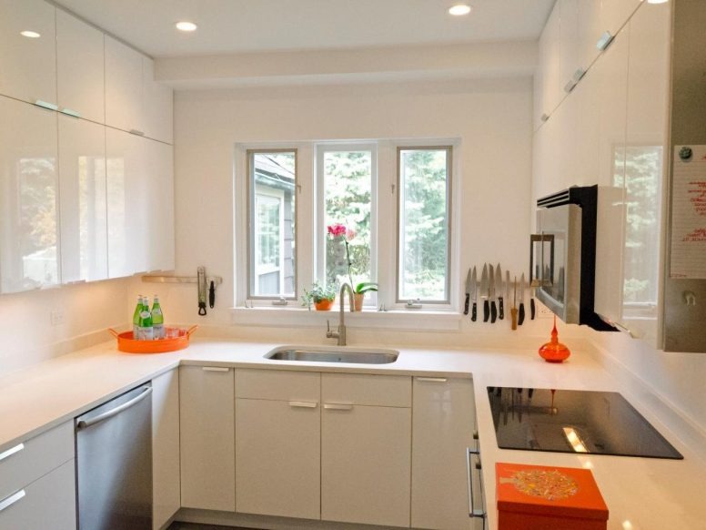 Nowoczesna kuchnia z białymi, lakierowanymi szafkami, jasnym blatem, oknem z widokiem na zieleń, akcentami w kolorze pomarańczowym i magnetycznym paskiem na noże.