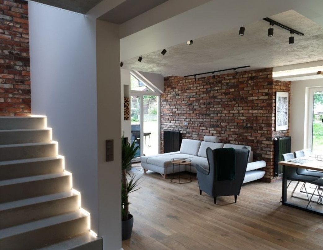 Nowoczesne wnętrze z ceglaną ścianą, drewnianą podłogą i podświetlanymi schodami, widok na salon z szarą sofą i fotelem.
