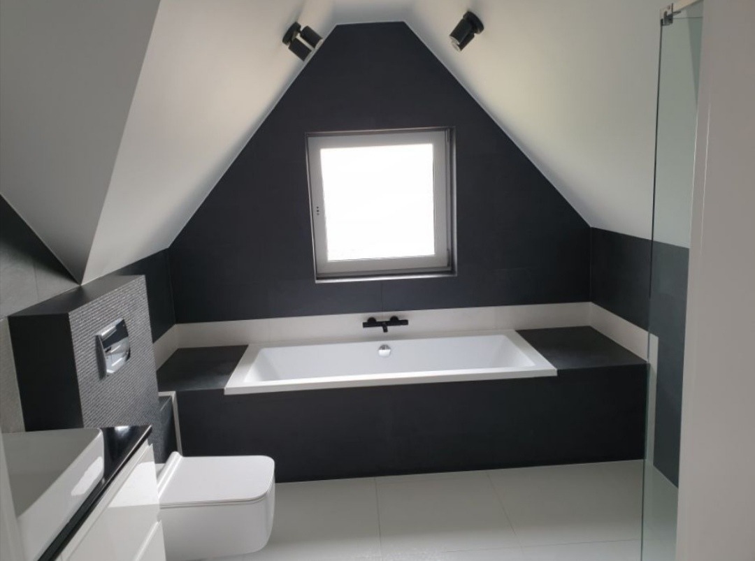 Nowoczesna łazienka na poddaszu z wanną w zabudowie, czarnymi ścianami i białymi płytkami podłogowymi, oświetlona światłem z okna dachowego i reflektorów.