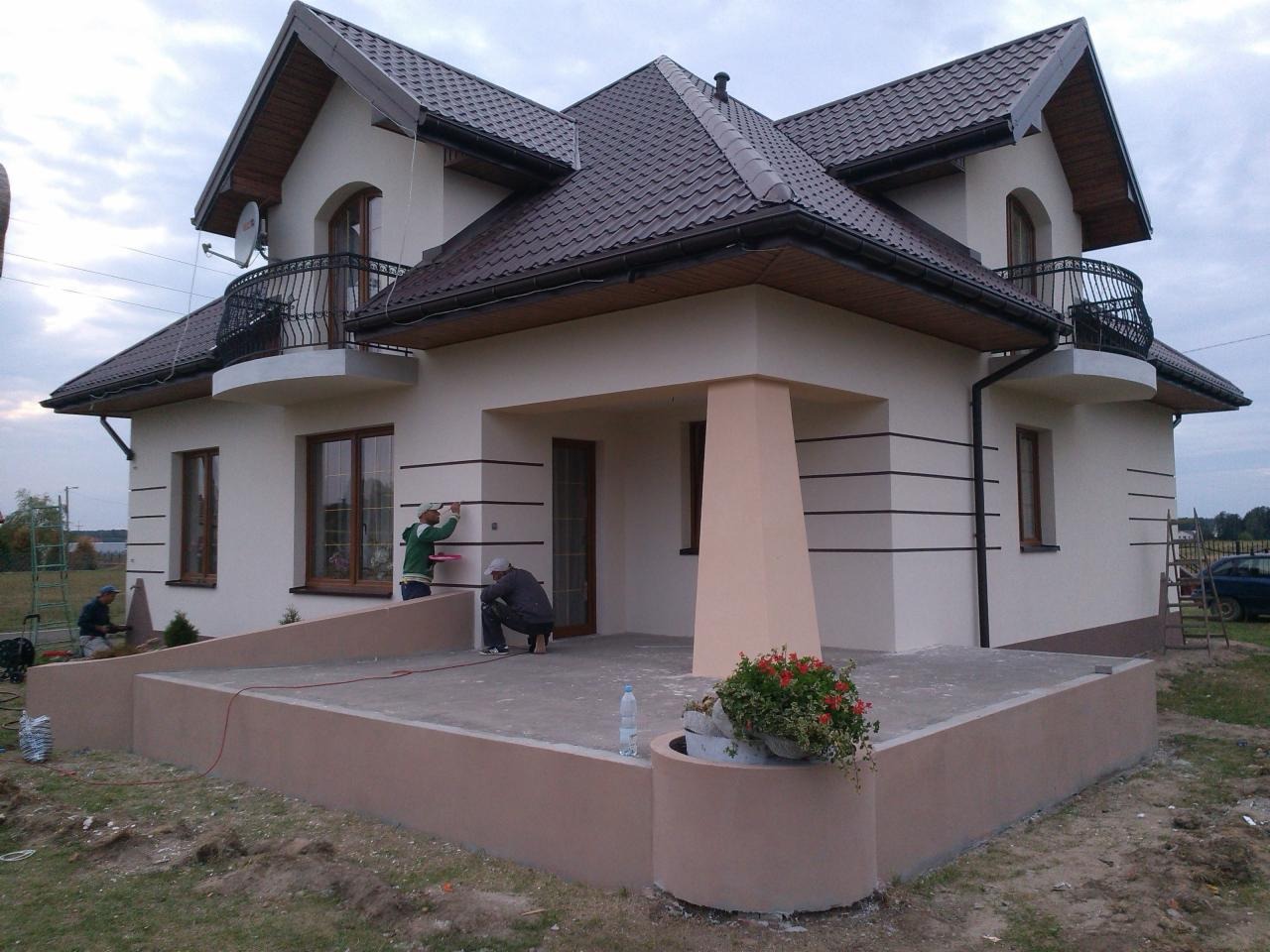 Wykończenie elewacji domu jednorodzinnego z brązowym dachem, widoczne detale w postaci poziomych pasów i pracujący przy elewacji robotnicy.