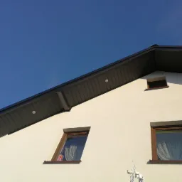 Fragment elewacji domu z ciemnoszarą blachodachówką, trzema oknami w brązowych ramach i błękitnym niebem w tle.