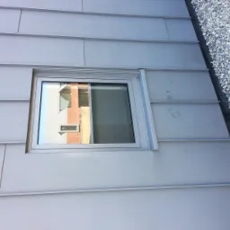 Szare blaszane poszycie dachu z oknem, w którym odbija się ceglany budynek, obok widoczne kamyki.