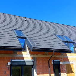 Firma ogólnobudowlana 'Deker' - Szary dach z dachówką ceramiczną, dwa okna dachowe, rynny w kolorze grafitowym, ściany budynku z czerwonej cegły dziurawki, błękitne niebo w tle.