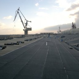Firma ogólnobudowlana 'Deker' - Rozległy, płaski dach pokryty papą termozgrzewalną w trakcie prac. W tle dźwigi portowe i pochmurne niebo. Widoczne rolki papy i ekipa montażowa.