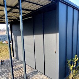 Firma ogólnobudowlana 'Deker' - Nowoczesna, grafitowa wiata garażowa z blachy trapezowej, widoczna zza metalowego ogrodzenia. Uwagę przykuwa minimalistyczny design i staranne wykonanie.