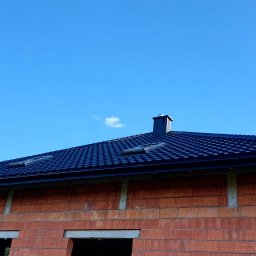 Firma ogólnobudowlana 'Deker' - Ciemna dachówka karpiówka na domu w stanie surowym otwartym, widoczne okna dachowe i komin na tle błękitnego nieba. Ujęcie z dołu, lekko z prawej strony.