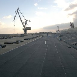 Firma ogólnobudowlana 'Deker' - Rozległy, płaski dach pokryty papą termozgrzewalną w trakcie prac. W tle dźwigi portowe i pochmurne niebo. Widoczne rolki papy i pracownicy.