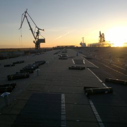 Firma ogólnobudowlana 'Deker' - Rozległy dach przemysłowy w trakcie hydroizolacji, z rolkami papy bitumicznej i kominkami wentylacyjnymi, na tle zachodzącego słońca i dźwigów portowych.