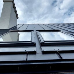 Firma ogólnobudowlana 'Deker' - Czarny dach z blachy trapezowej z dwoma oknami dachowymi, widok z dołu na tle pochmurnego nieba i komina. Fragment dachu z drewnianą konstrukcją w tle.