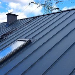 Firma ogólnobudowlana 'Deker' - Szary dach z blachy trapezowej z trzema oknami dachowymi i kominem na tle błękitnego nieba z chmurami i słupem wysokiego napięcia w oddali.