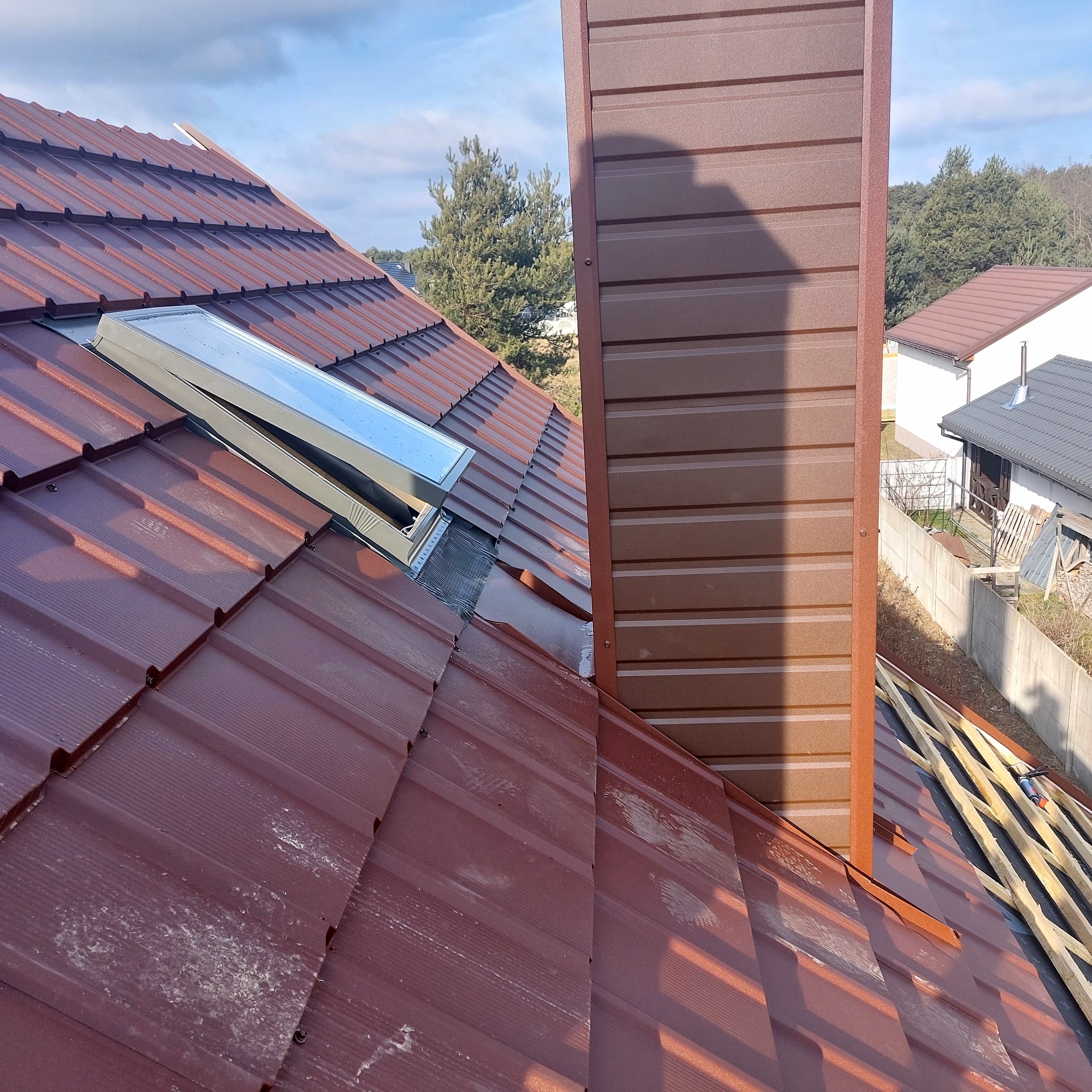 Ciemnobrązowy dach z blachy trapezowej z oknem dachowym i kominem. Widoczne elementy konstrukcji dachu. Częściowo zabrudzona powierzchnia blachy.