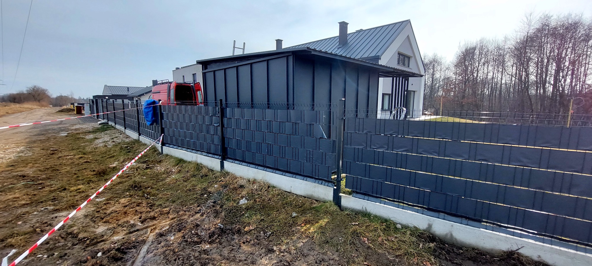 Nowoczesny, grafitowy płot panelowy z podmurówką betonową, na tle domu z dachem z blachy w tym samym kolorze. Czerwony samochód dostawczy w tle.