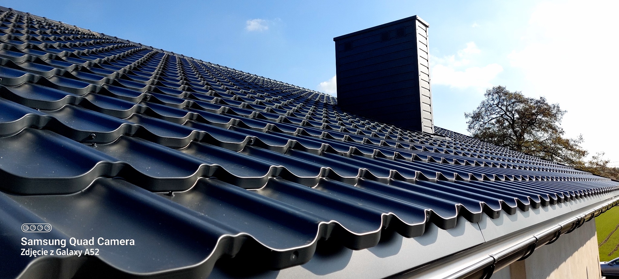 Nowy, czarny dach z dachówki modułowej, z widocznym kominem i rynną. Ujęcie z dołu, podkreślające regularność wzoru i estetykę wykonania. Błękitne niebo w tle.