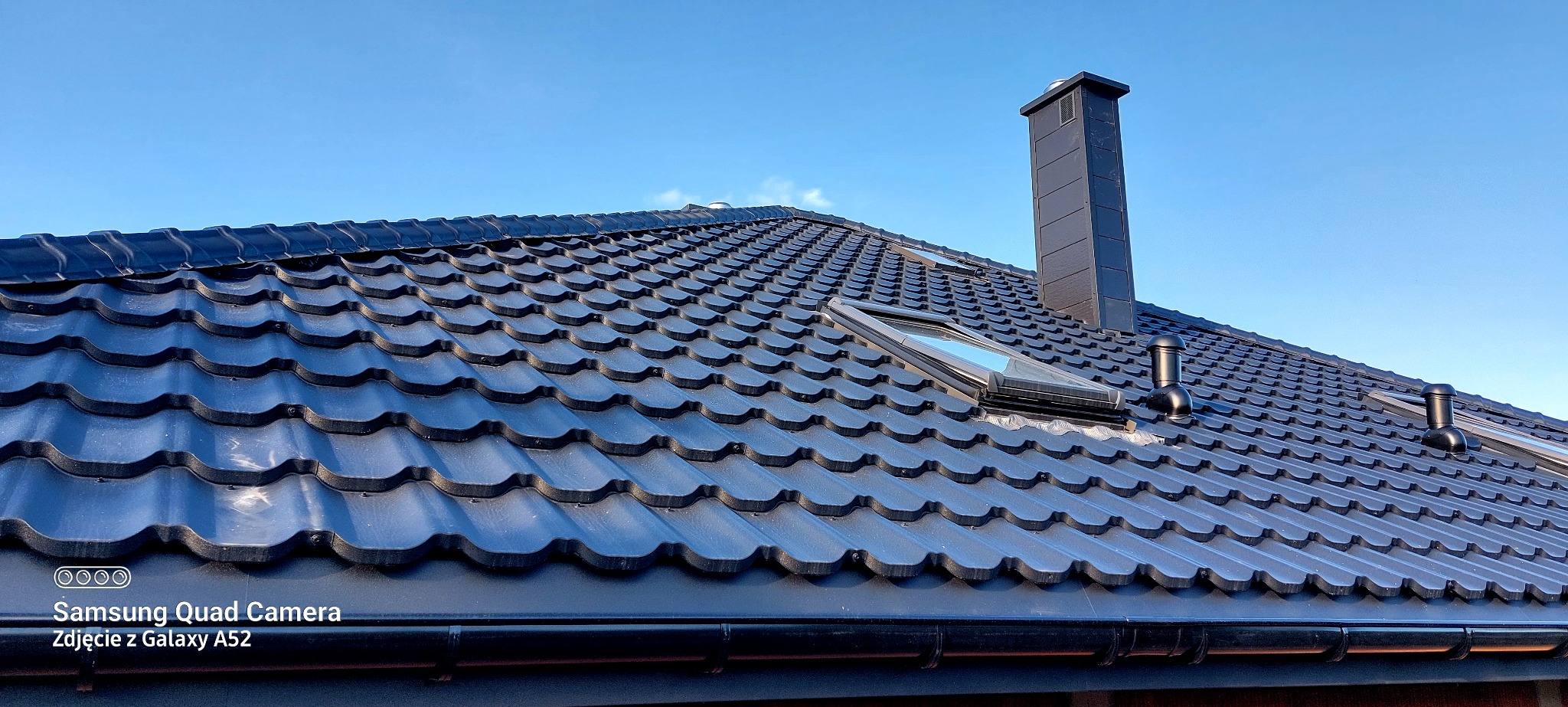 Czarny dach z dachówki modułowej, z oknem dachowym i kominem na tle błękitnego nieba. Widoczne rynny i odpowietrzniki. Ujęcie z dołu, podkreślające geometrię pokrycia.