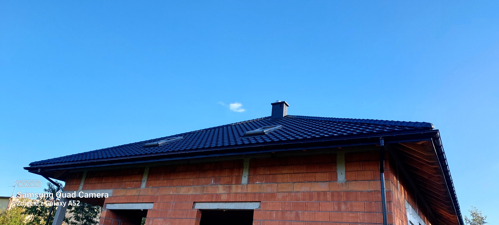 Ciemna dachówka karpiówka na domu w stanie surowym otwartym, widoczne okna dachowe i komin na tle błękitnego nieba. Ujęcie z dołu, lekko z prawej strony.