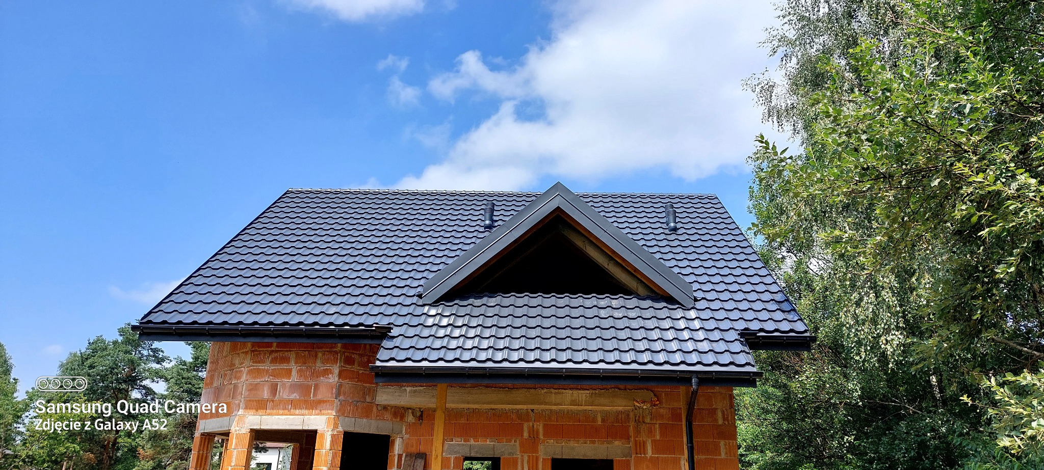 Nowy, grafitowy dach z dachówki ceramicznej na surowym budynku z czerwonej cegły, widok z dołu na tle błękitnego nieba z chmurami i zieleni drzew.