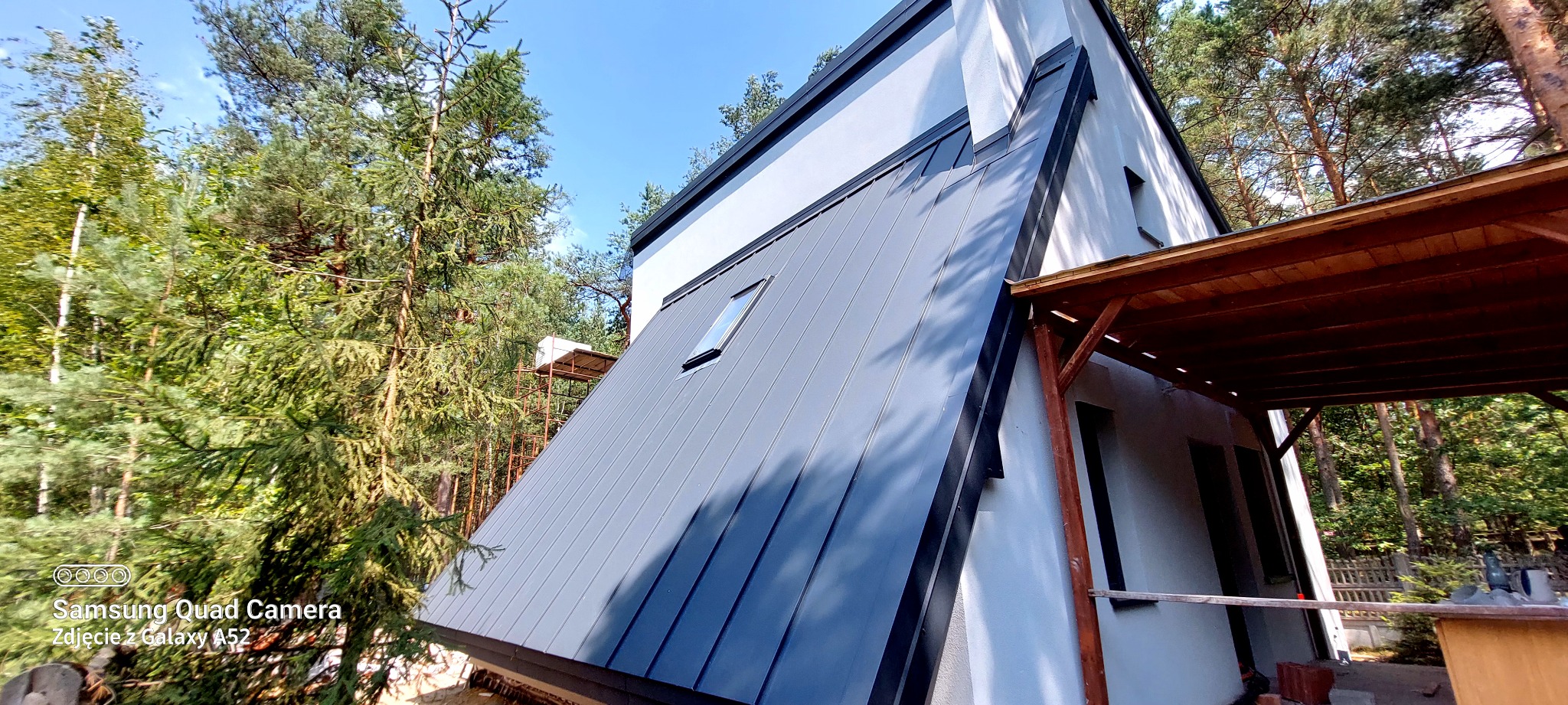 Nowoczesny dom z szarą blachą na dachu i oknem połaciowym, otoczony zielenią. Widok z dołu, podkreślający geometrię dachu i kontrast z otoczeniem.